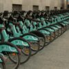 Row of turquoise Bizî bike-sharing bicycles lined up along a city sidewalk near a stone building façade.