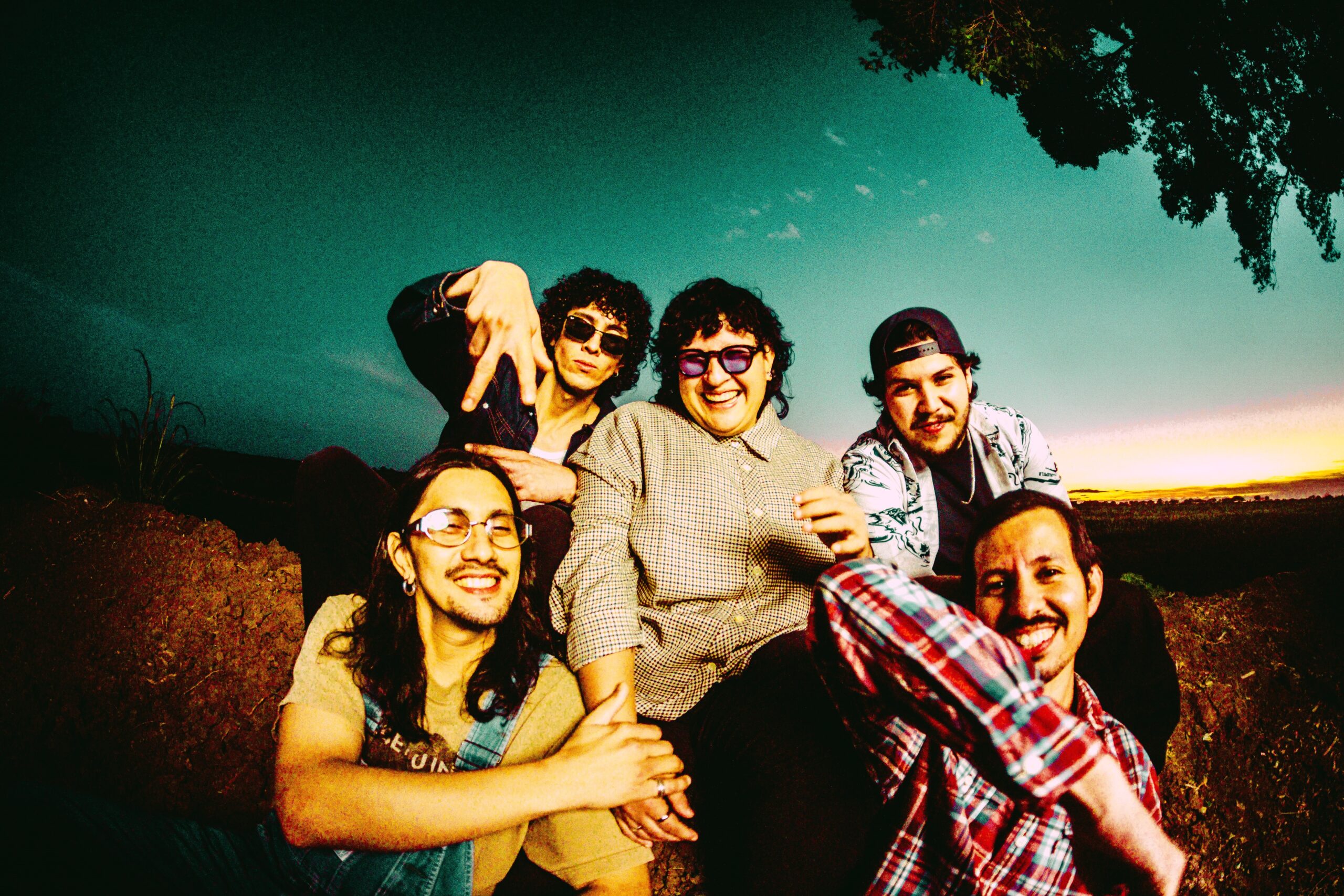 Group of friends posing closely for a selfie outdoors at sunset, smiling and making playful gestures toward the camera.