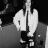 Black-and-white photo of a person wearing headphones, a striped shirt, a black tie, and a patchwork skirt, standing by a wall with cords on the floor.