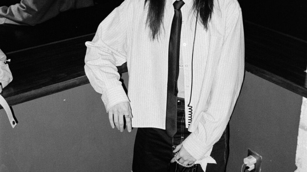 Black-and-white photo of a person wearing headphones, a striped shirt, a black tie, and a patchwork skirt, standing by a wall with cords on the floor.