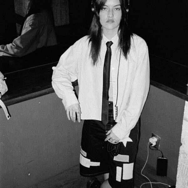 Black-and-white photo of a person wearing headphones, a striped shirt, a black tie, and a patchwork skirt, standing by a wall with cords on the floor.