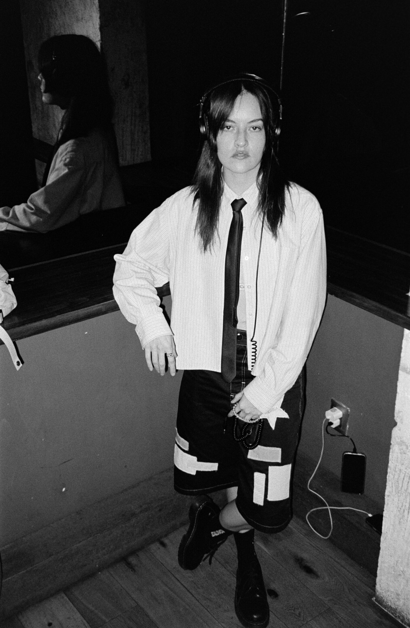 Black-and-white photo of a person wearing headphones, a striped shirt, a black tie, and a patchwork skirt, standing by a wall with cords on the floor.