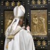 Catholic bishop in white ceremonial robes standing before a large ornate door with gold relief panels depicting biblical scenes
