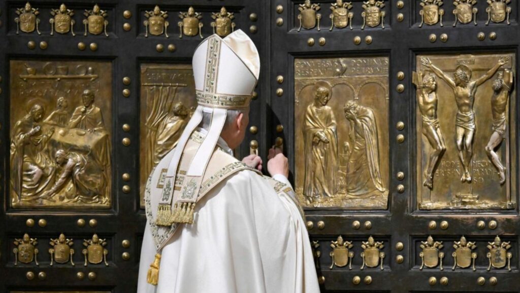 Catholic bishop in white ceremonial robes standing before a large ornate door with gold relief panels depicting biblical scenes