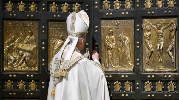 Catholic bishop in white ceremonial robes standing before a large ornate door with gold relief panels depicting biblical scenes