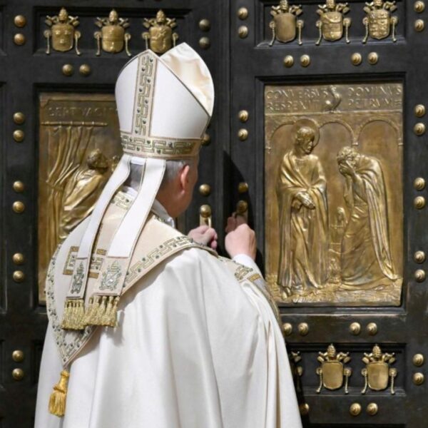 Catholic bishop in white ceremonial robes standing before a large ornate door with gold relief panels depicting biblical scenes