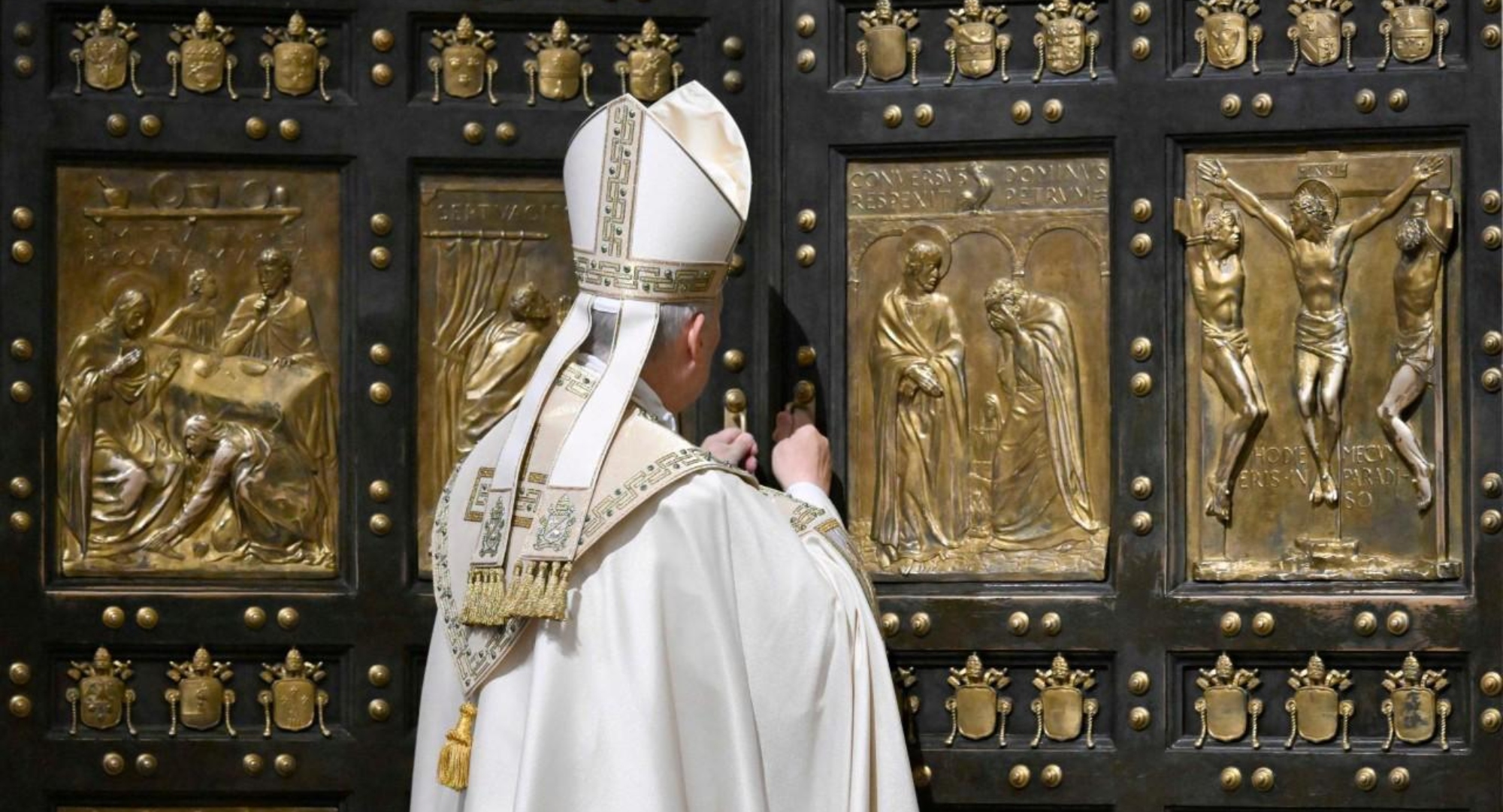 Catholic bishop in white ceremonial robes standing before a large ornate door with gold relief panels depicting biblical scenes