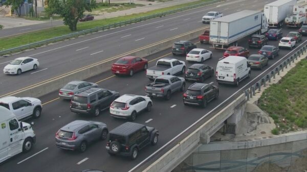 Heavy traffic on a multi-lane highway with cars and trucks lined up in each direction, separated by a concrete barrier and an overpass on the right.