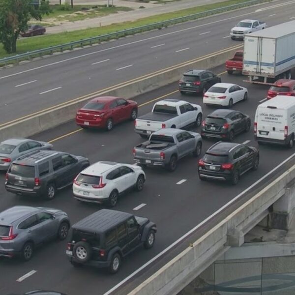 Heavy traffic on a multi-lane highway with cars and trucks lined up in each direction, separated by a concrete barrier and an overpass on the right.