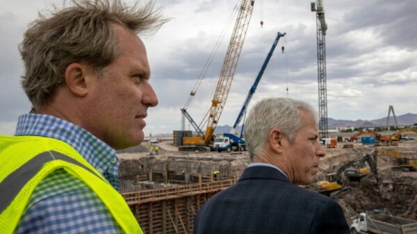 Two men overlook a construction site with cranes and heavy machinery in a desert landscape beneath a cloudy sky, one wearing a neon safety vest and the other in a suit.