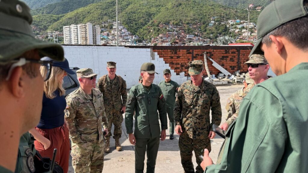 Group of soldiers in uniform standing in a circle and talking in a ruined outdoor area with hills in the background.