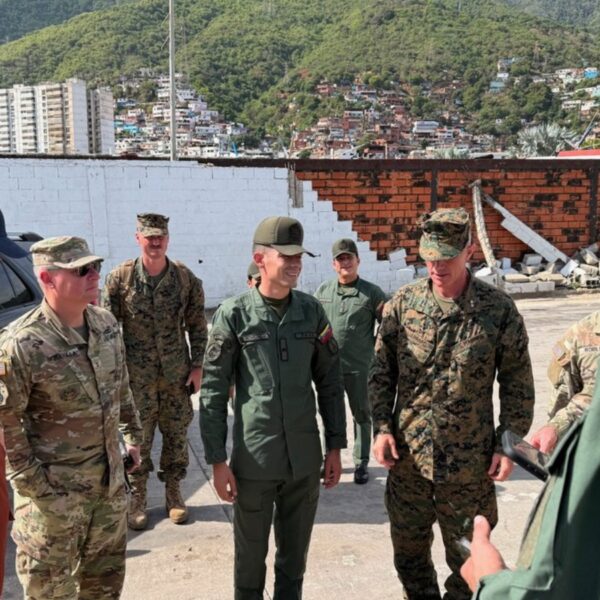 Group of soldiers in uniform standing in a circle and talking in a ruined outdoor area with hills in the background.