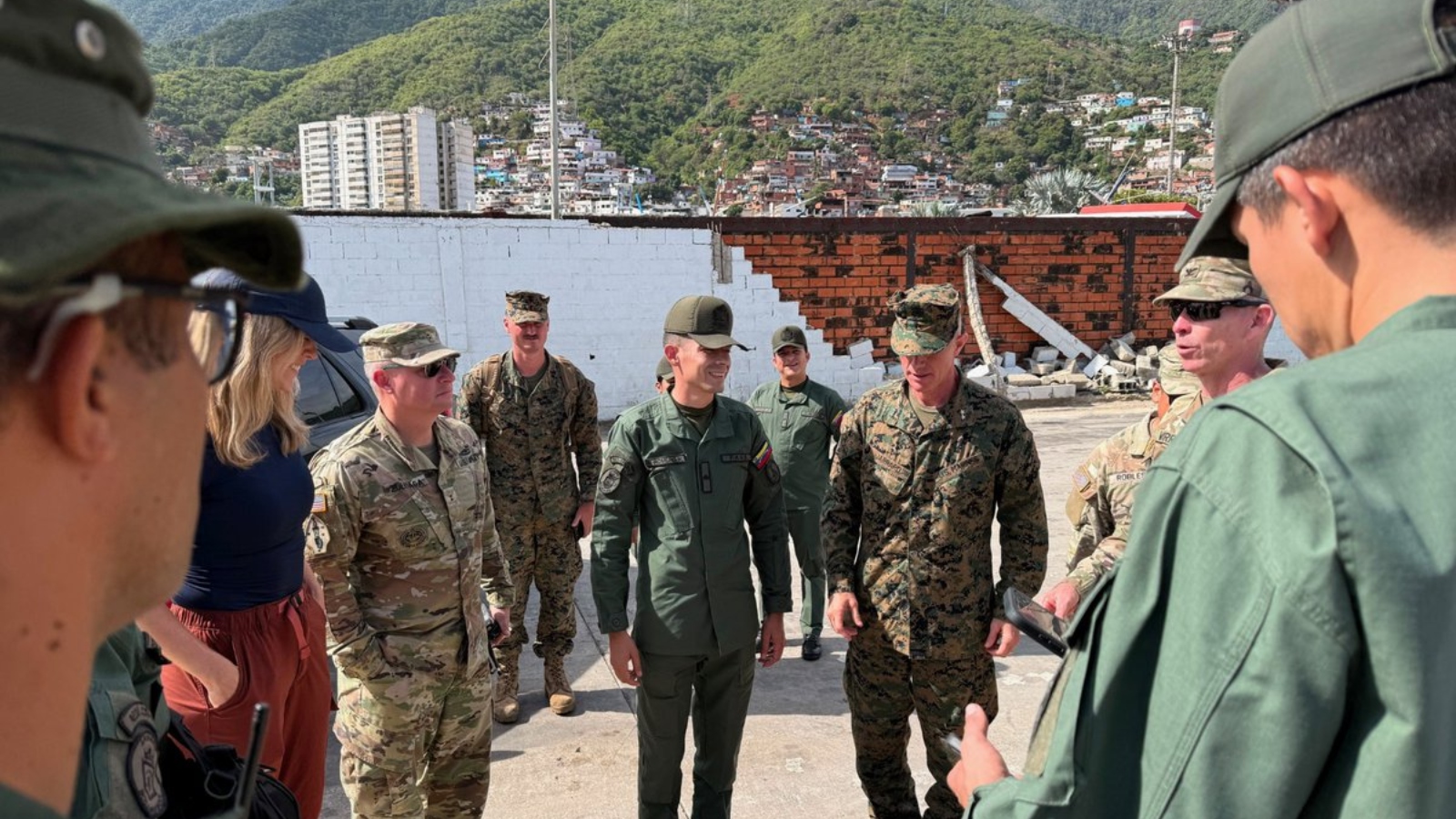 Group of soldiers in uniform standing in a circle and talking in a ruined outdoor area with hills in the background.