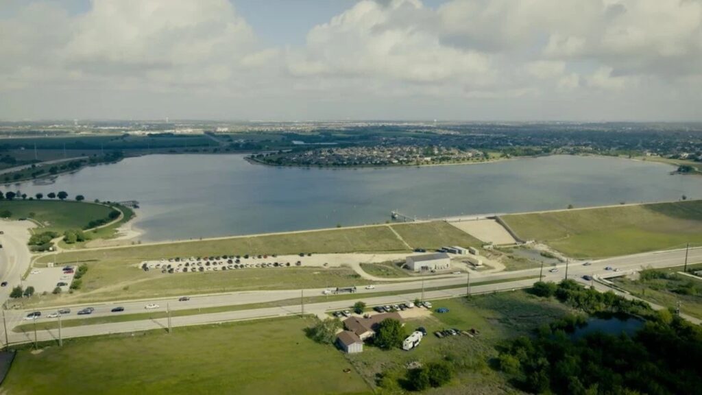 Aerial view of a large lake with a grassy shoreline, park areas, and a road along the edge.