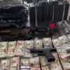 Rifle and a handgun lie on a table amid large stacks of cash, ammo boxes, and assorted gear on a cluttered surface.