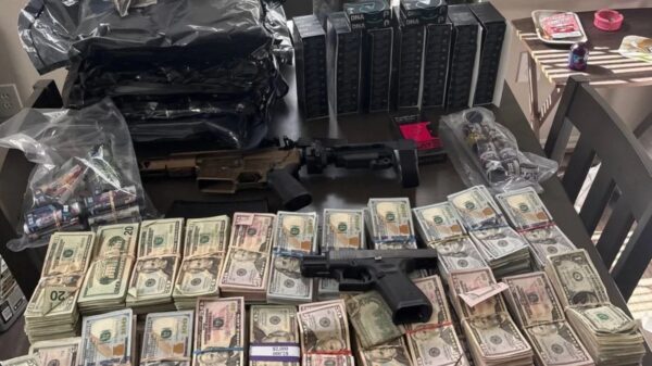 Rifle and a handgun lie on a table amid large stacks of cash, ammo boxes, and assorted gear on a cluttered surface.