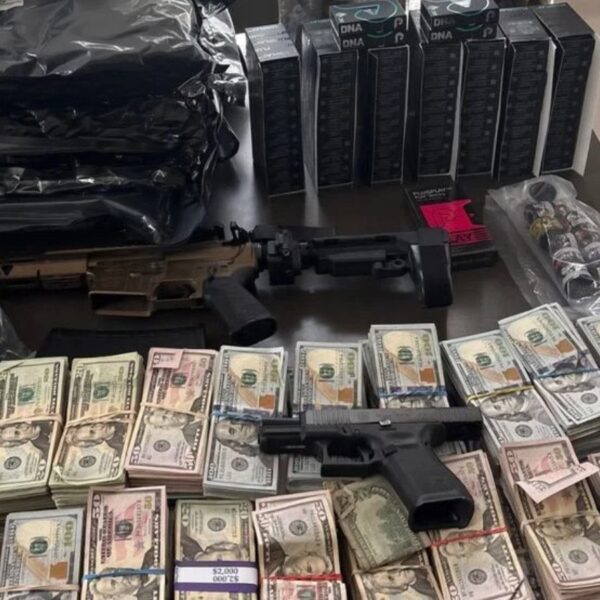 Rifle and a handgun lie on a table amid large stacks of cash, ammo boxes, and assorted gear on a cluttered surface.