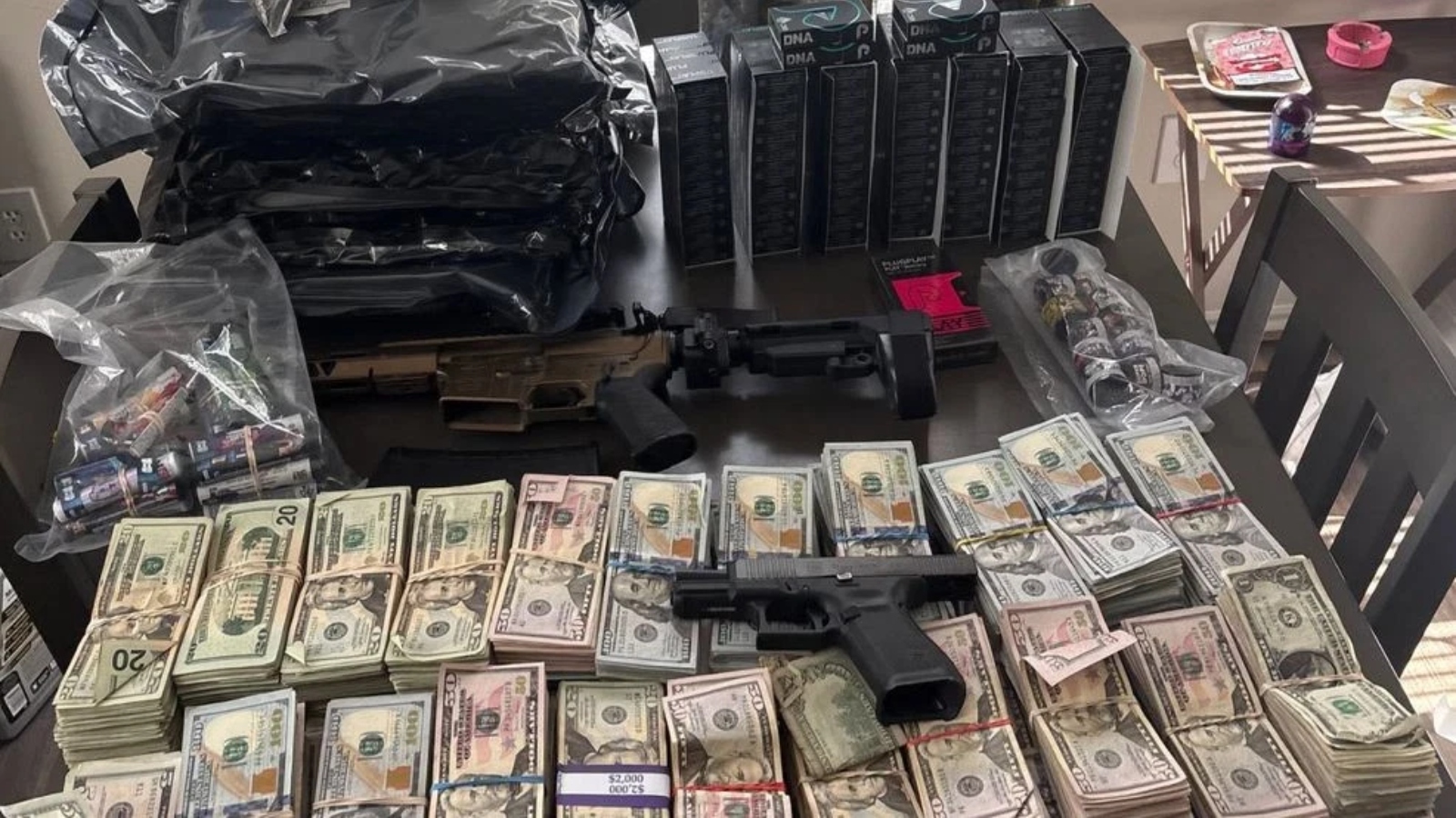 Rifle and a handgun lie on a table amid large stacks of cash, ammo boxes, and assorted gear on a cluttered surface.