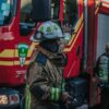 Firefighters in full turnout gear near a red fire truck with reflective stripes, one raises a hand as they prepare equipment.