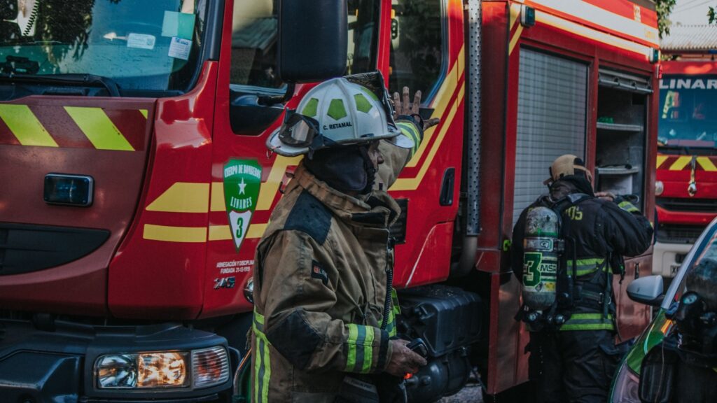 Firefighters in full turnout gear near a red fire truck with reflective stripes, one raises a hand as they prepare equipment.