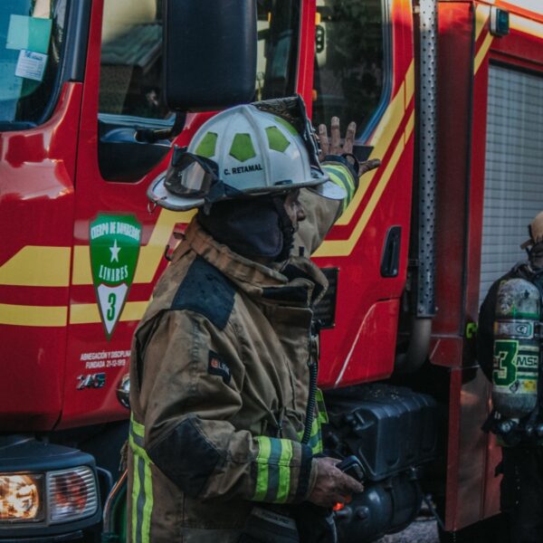 Firefighters in full turnout gear near a red fire truck with reflective stripes, one raises a hand as they prepare equipment.