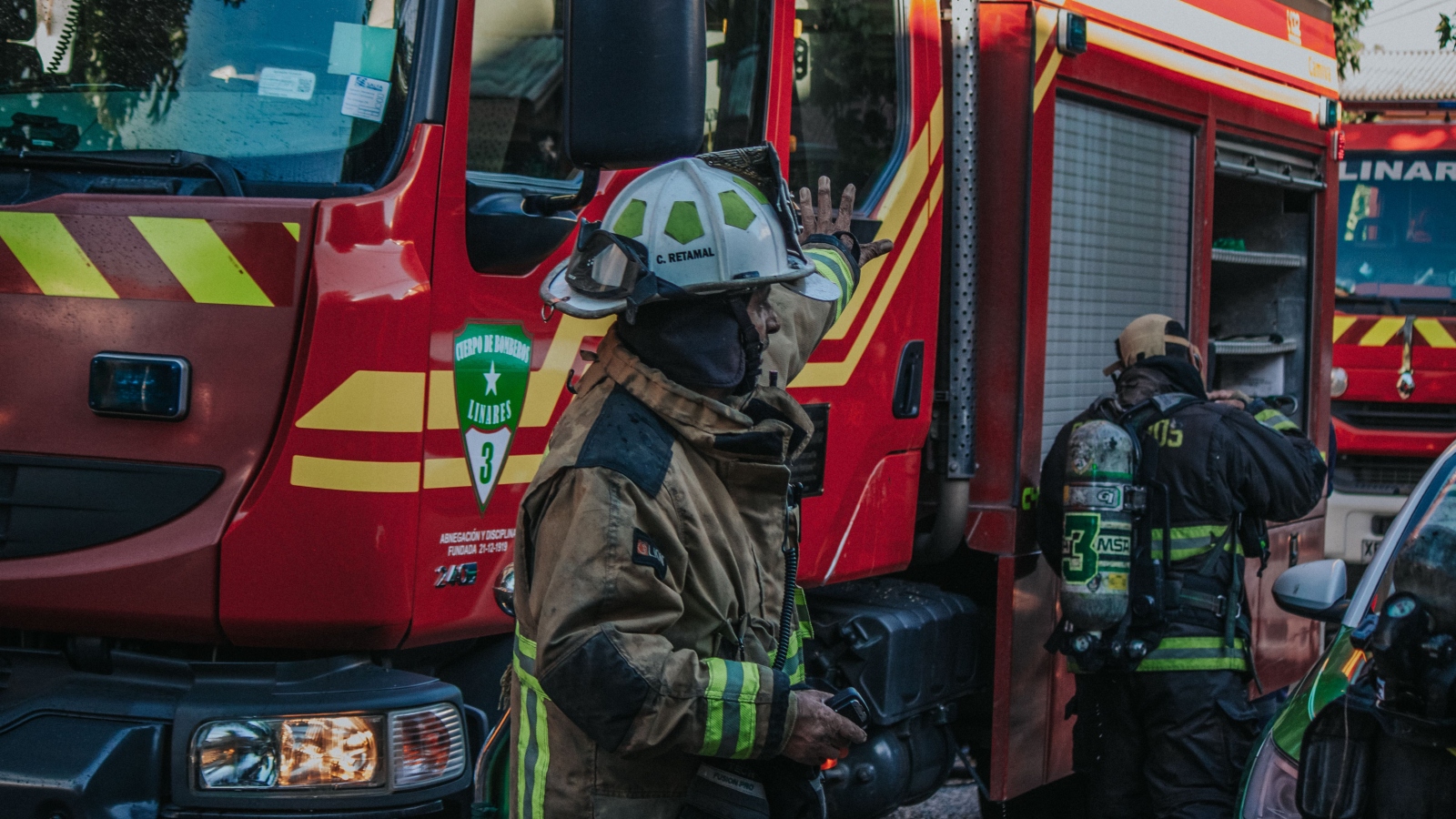 Firefighters in full turnout gear near a red fire truck with reflective stripes, one raises a hand as they prepare equipment.