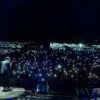 A concert stage at night with two performers facing a massive crowd, each audience member holding up lit phones to create a twinkling sea of lights.