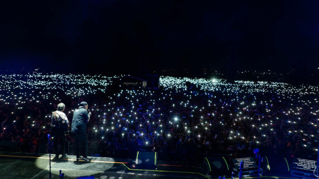 A concert stage at night with two performers facing a massive crowd, each audience member holding up lit phones to create a twinkling sea of lights.