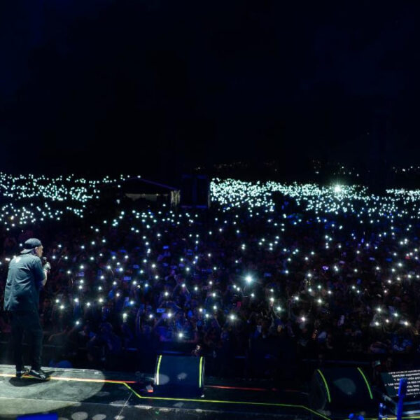 A concert stage at night with two performers facing a massive crowd, each audience member holding up lit phones to create a twinkling sea of lights.