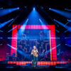 A singer performs center stage under blue spotlights with a large, red-framed screen behind showing abstract drawings.
