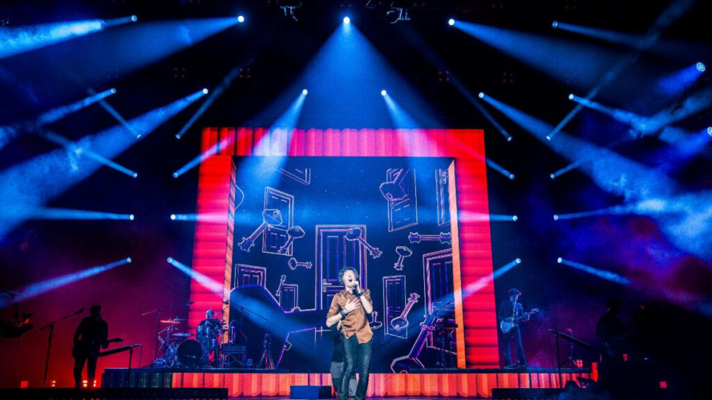 A singer performs center stage under blue spotlights with a large, red-framed screen behind showing abstract drawings.