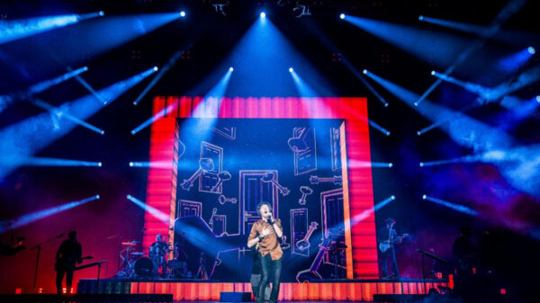 A singer performs center stage under blue spotlights with a large, red-framed screen behind showing abstract drawings.