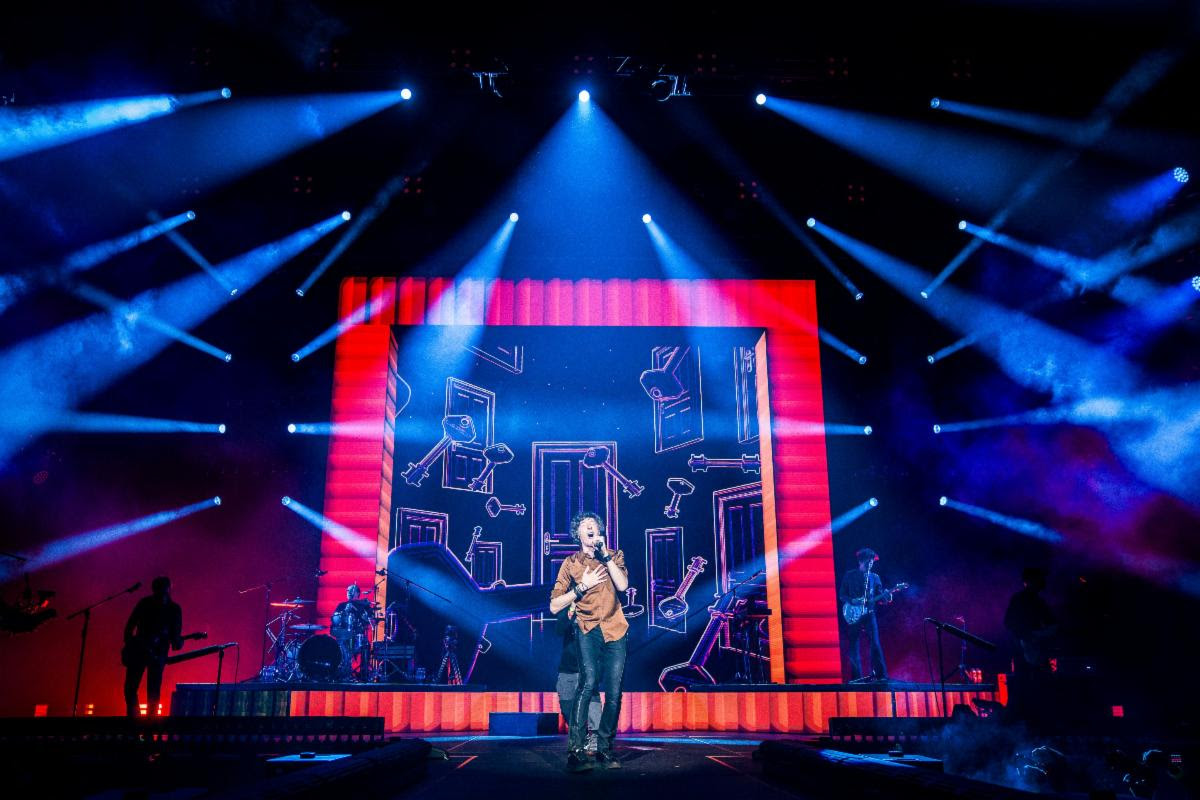 A singer performs center stage under blue spotlights with a large, red-framed screen behind showing abstract drawings.