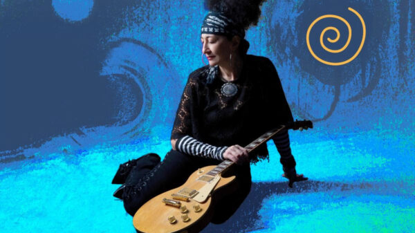 Musician sitting with a gold electric guitar against a blue abstract background with an orange spiral logo on the right.