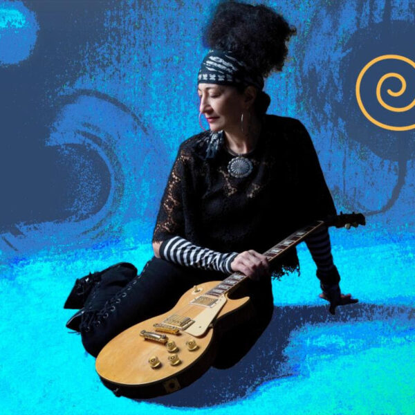 Musician sitting with a gold electric guitar against a blue abstract background with an orange spiral logo on the right.