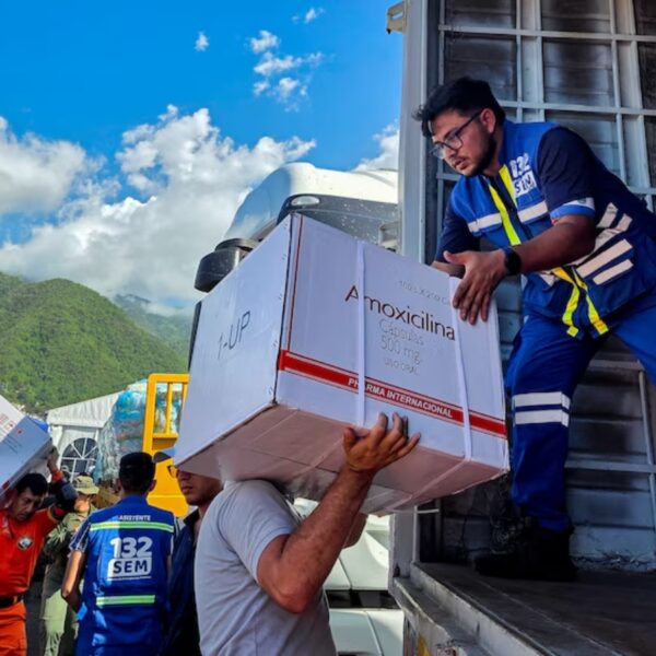 Relief workers unload a large medicine box labeled 'Amoxicillina' from a truck during disaster relief. Mountain backdrop and blue sky.