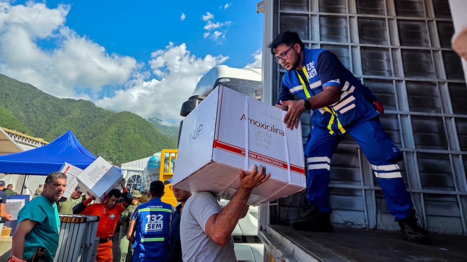 Relief workers unload a large medicine box labeled 'Amoxicillina' from a truck during disaster relief. Mountain backdrop and blue sky.