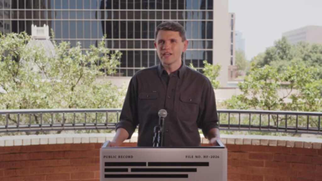 Man speaks at an outdoor podium with a microphone, city buildings and trees in the background.