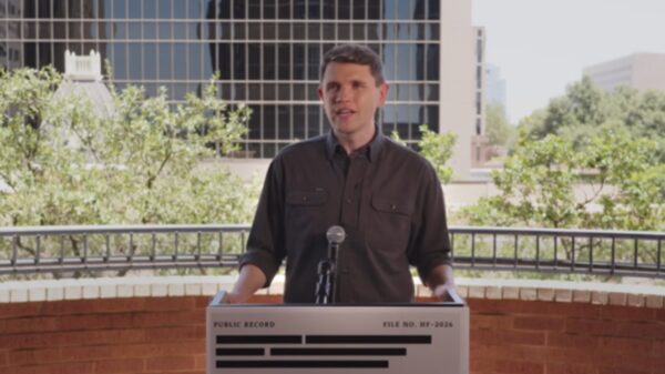 Man speaks at an outdoor podium with a microphone, city buildings and trees in the background.