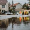 Workers in yellow rain gear filling sandbags to block floodwater on a residential street with a car nearby.
