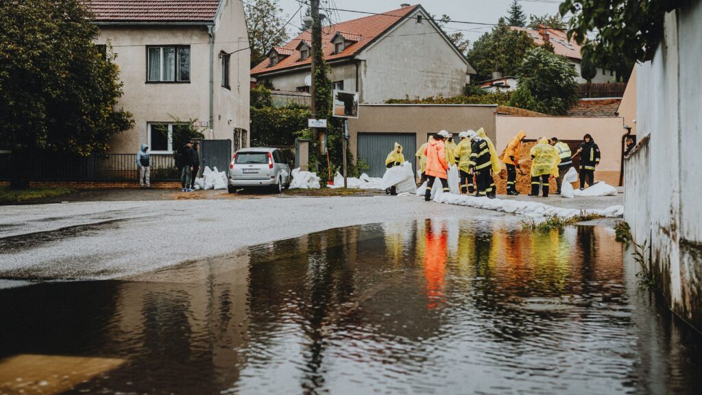 Workers in yellow rain gear filling sandbags to block floodwater on a residential street with a car nearby.