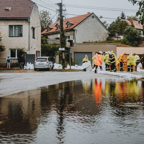 Workers in yellow rain gear filling sandbags to block floodwater on a residential street with a car nearby.