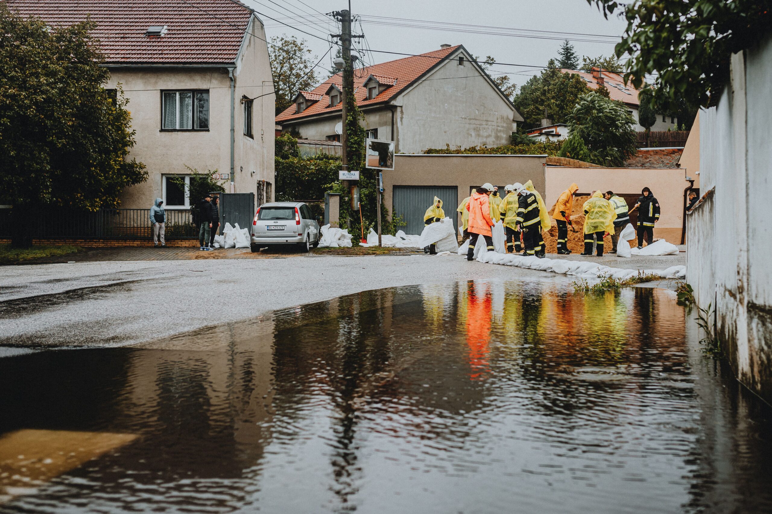 Workers in yellow rain gear filling sandbags to block floodwater on a residential street with a car nearby.