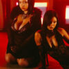 Two women in black lace outfits kneeling on a red-lit stage, one with short dark hair and fishnet stockings, the other with long dark hair; both wearing provocative attire.