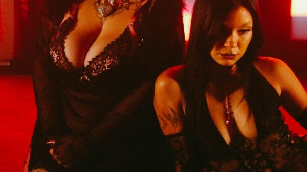 Two women in black lace outfits kneeling on a red-lit stage, one with short dark hair and fishnet stockings, the other with long dark hair; both wearing provocative attire.