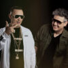 Two men wearing sunglasses pose together in a studio; the man on the left wears a white jacket and gold chains, the man on the right in a dark green jacket.
