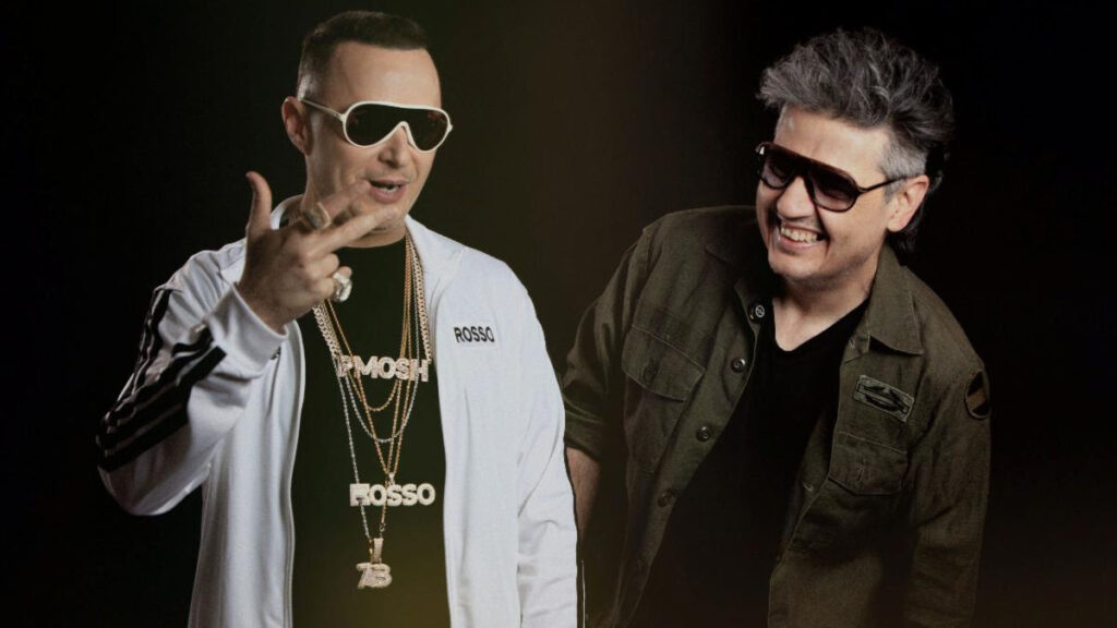 Two men wearing sunglasses pose together in a studio; the man on the left wears a white jacket and gold chains, the man on the right in a dark green jacket.