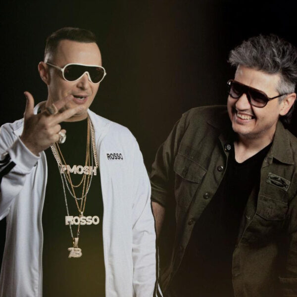 Two men wearing sunglasses pose together in a studio; the man on the left wears a white jacket and gold chains, the man on the right in a dark green jacket.