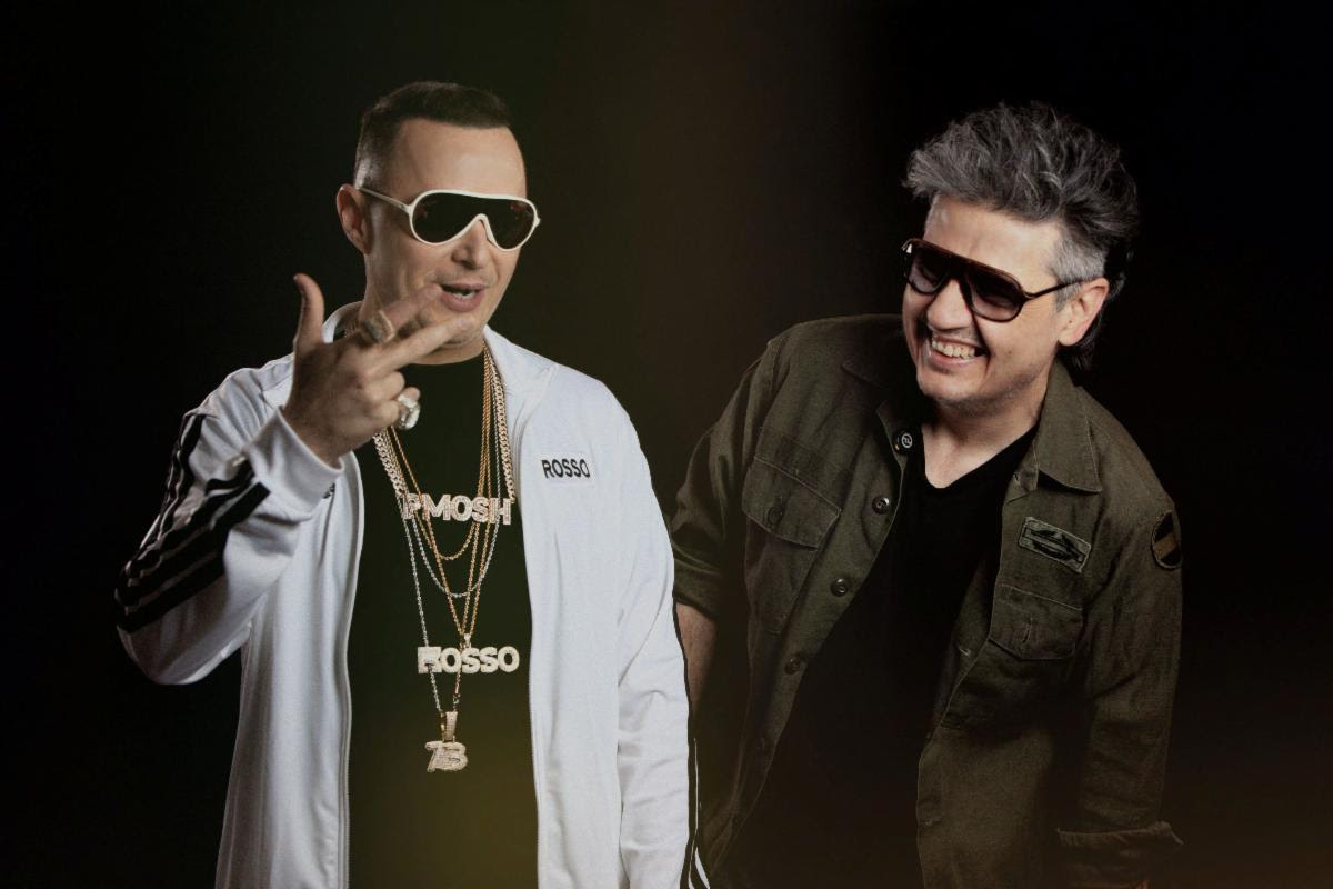 Two men wearing sunglasses pose together in a studio; the man on the left wears a white jacket and gold chains, the man on the right in a dark green jacket.