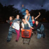 Six cheerful performers in colorful patched outfits pose around a red shopping cart on a nighttime street, arms raised.